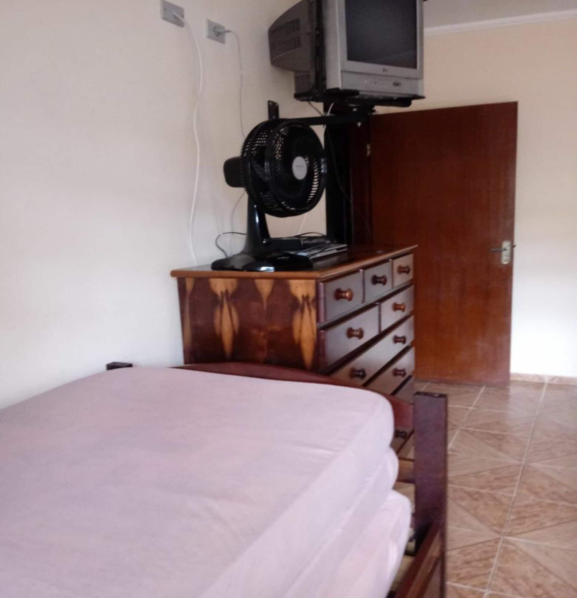 Um quarto com uma cama, cômoda, ventoinha e televisão sobre um móvel.