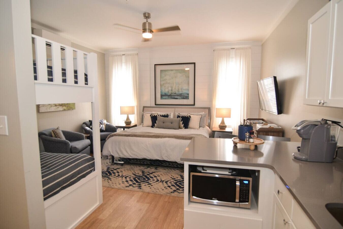 Une chambre d’hôtel moderne équipée d’un lit, de tables de chevet, d’un bureau avec un micro-ondes, d’un ventilateur de plafond et d’une grande fenêtre avec des rideaux.