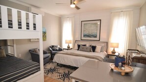 Una habitación de hotel moderna con una cama litera, una cama individual, un escritorio y una silla. Hay un ventilador de techo, un cuadro en la pared y una ventana con cortinas.