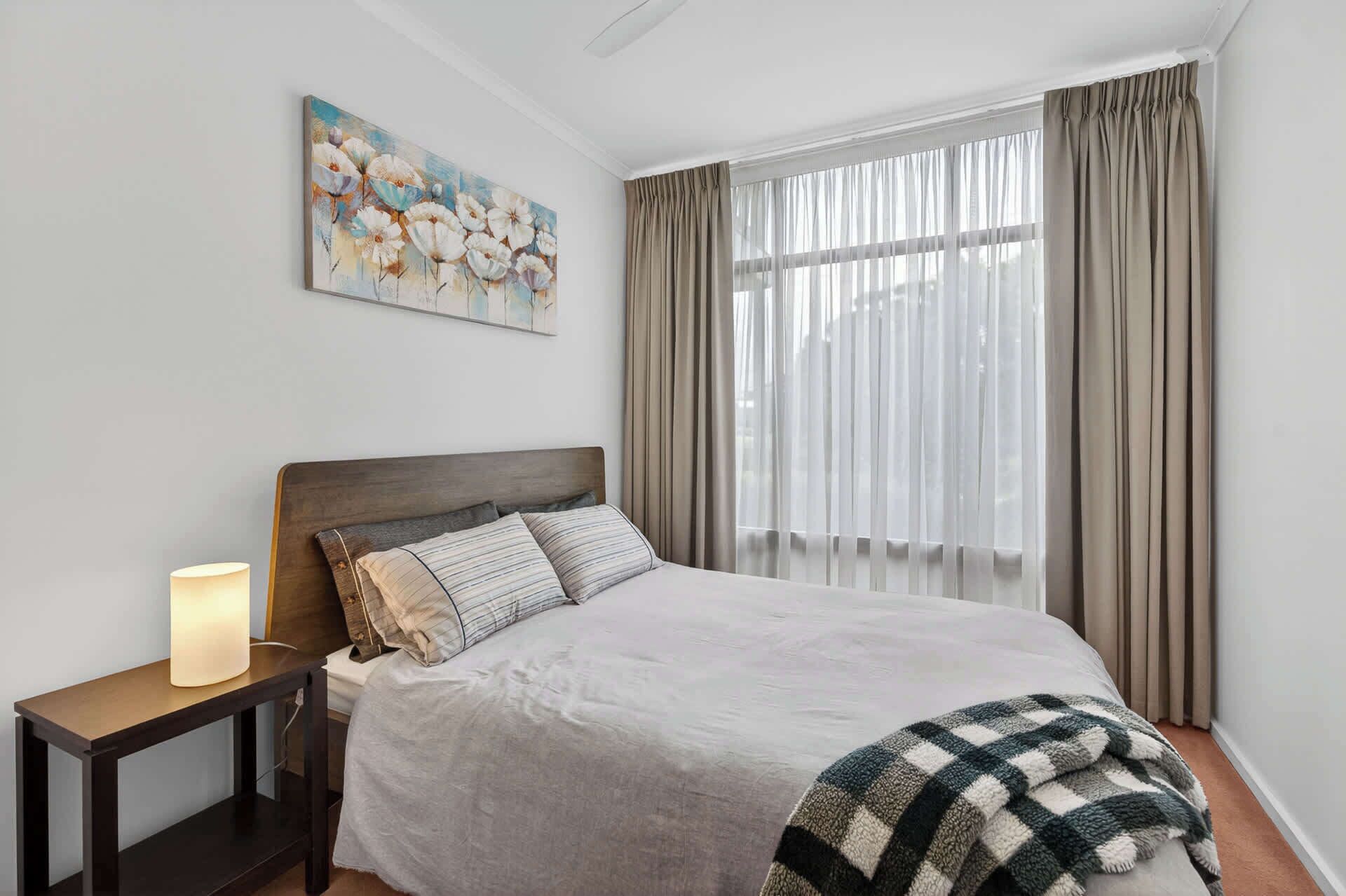 Un dormitorio con cama, lámpara de noche, un cuadro y cortinas traslúcidas.