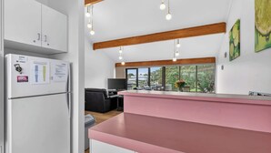A modern kitchen with a pink countertop, white cabinets, and a built-in refrigerator. There is a dining area with a black sofa and a TV. Large windows provide a view of the outdoors. - 4/1 Harbour View Terrace - Wi-Fi (Victor Harbor)