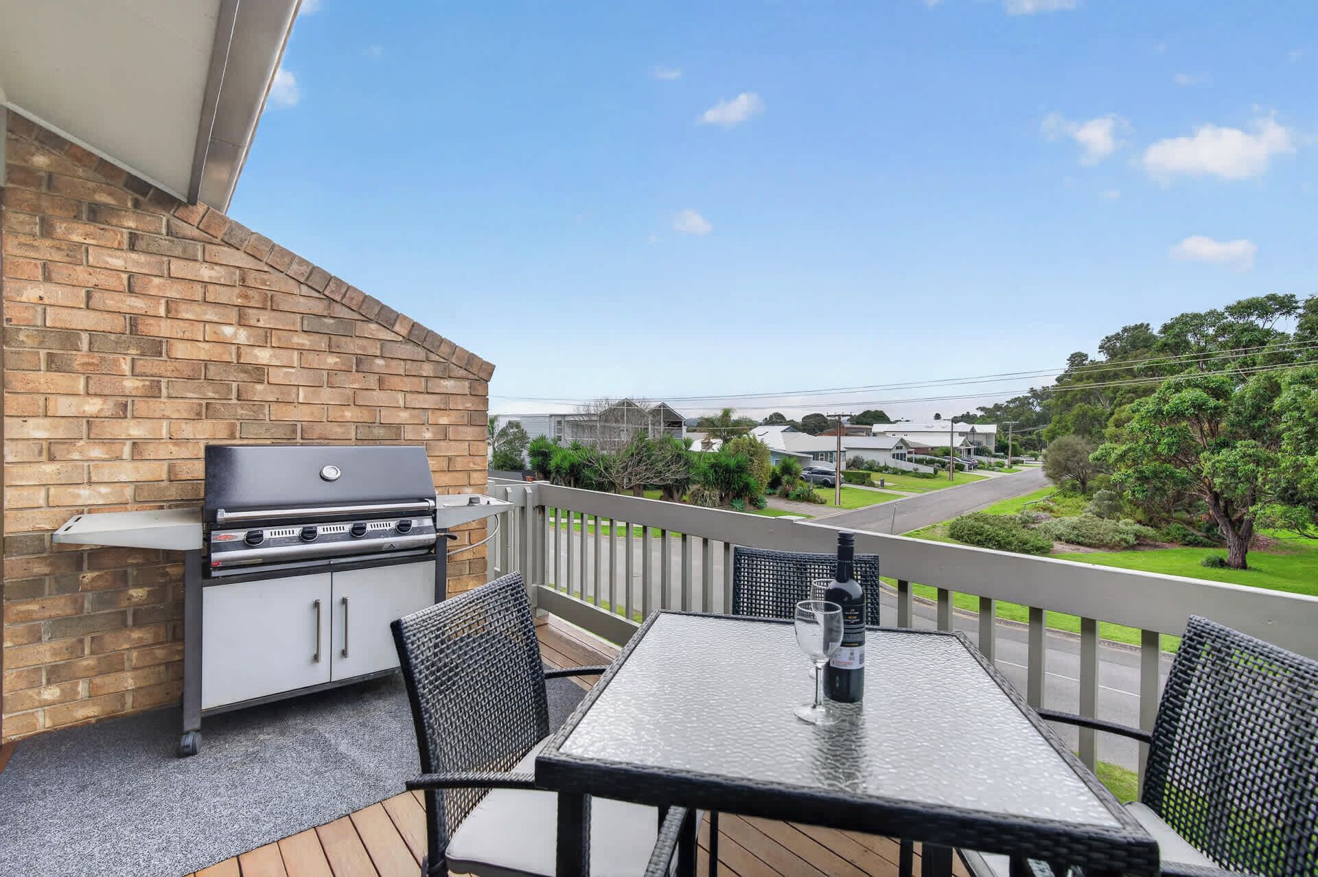 A balcony with a grill, a table, and chairs, overlooking a street and greenery.