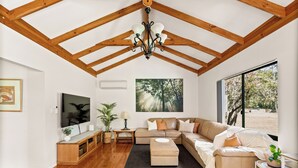 A spacious living room with a high ceiling, wooden beams, a large window, a sectional sofa, a TV, a plant, and a fireplace.