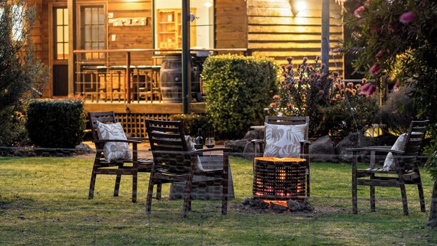 A cozy outdoor seating area with wooden chairs and a fire pit, surrounded by a well-maintained garden and a wooden building with string lights.