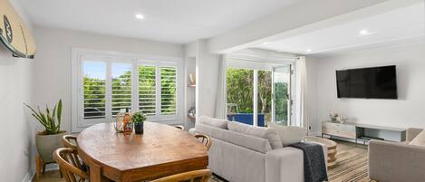 A modern living room with a dining area, a sofa, a TV, and large windows.