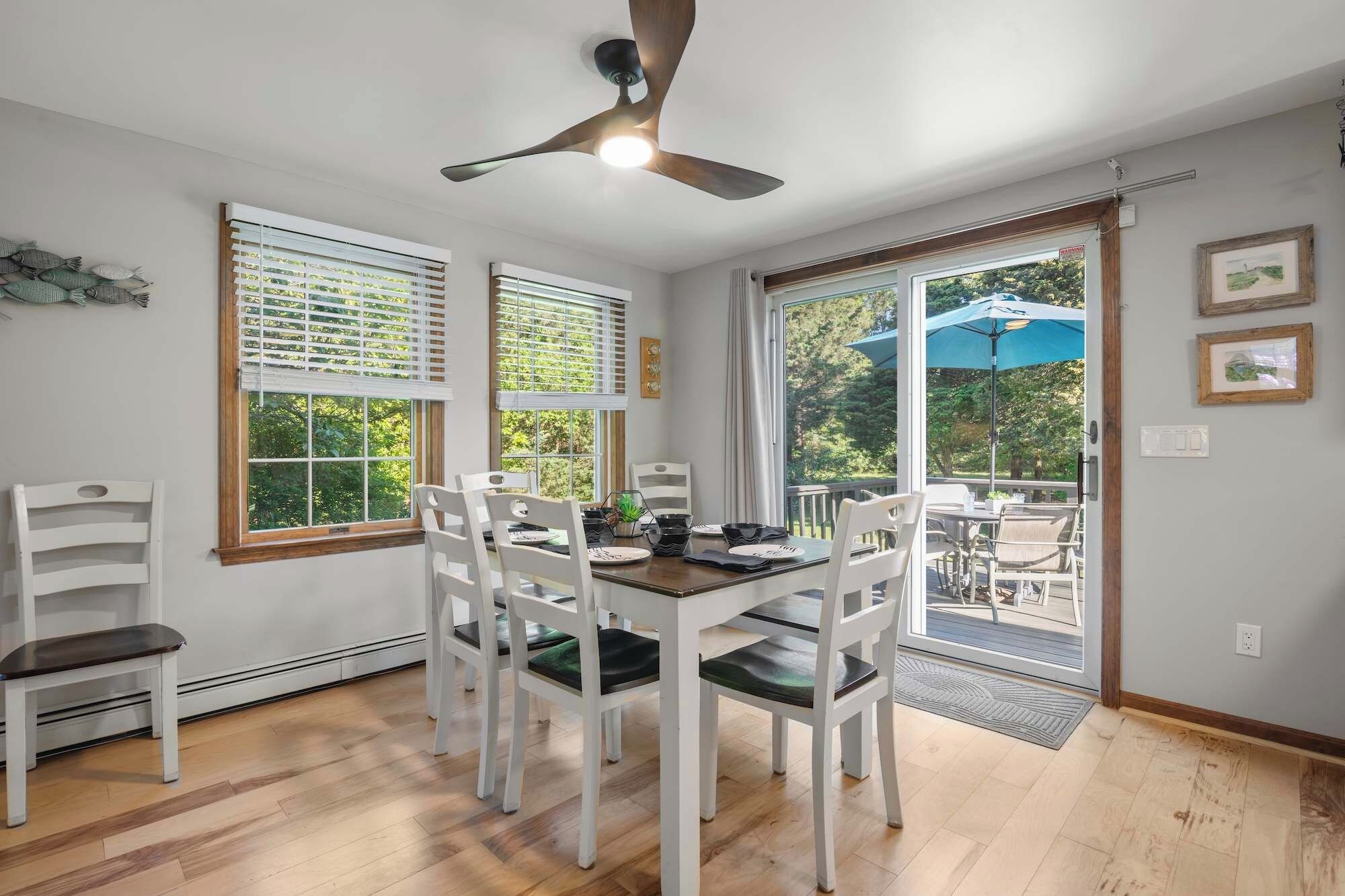A dining area with a table set for four, white chairs, a ceiling fan, and large windows with blinds.