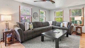 A living room with a sectional sofa, a coffee table, a ceiling fan, and large windows with blinds. - Perfect Relaxation Spot 5 min to Beach! (Eastham)