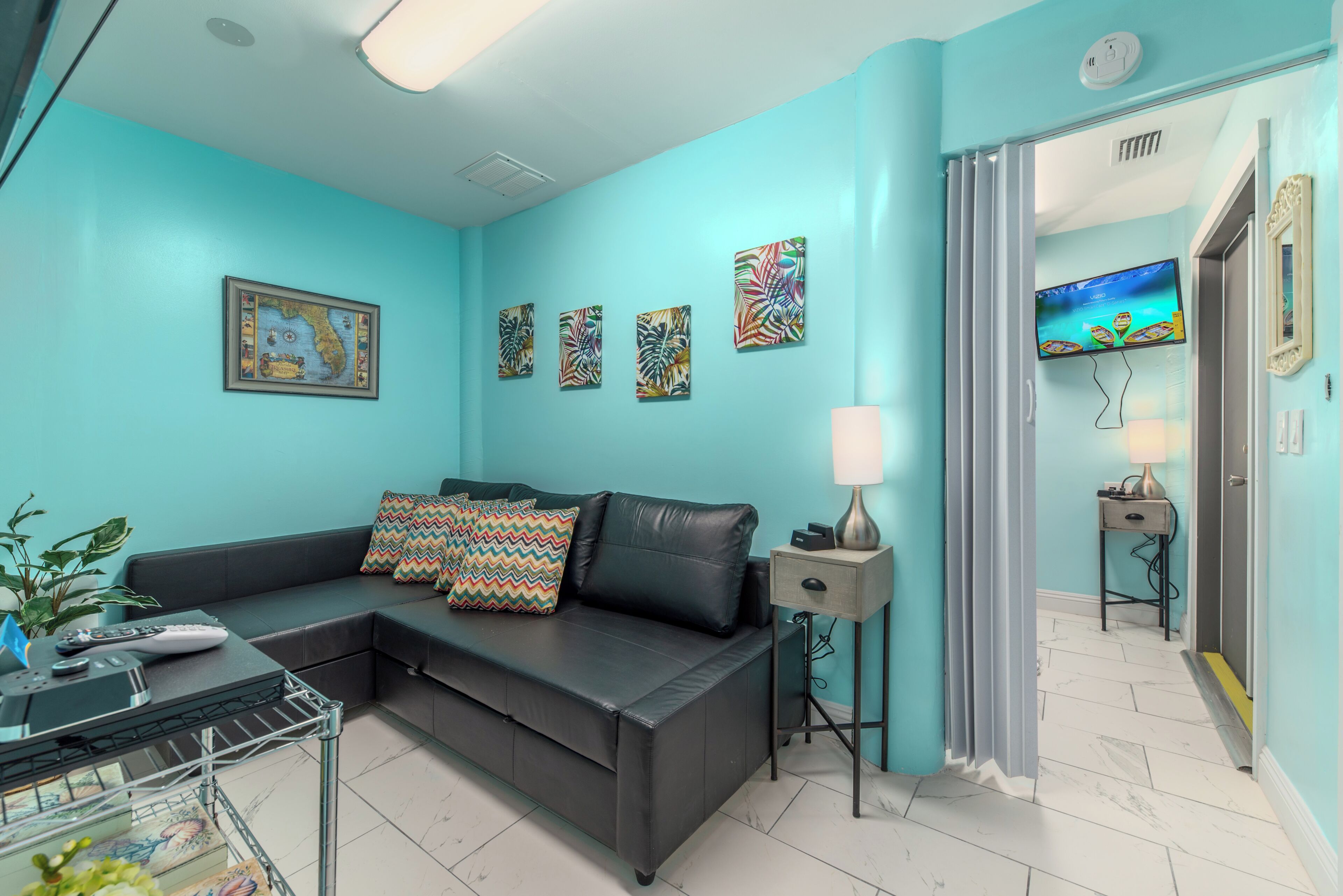 Een woonkamer met een turquoise wand, een zwarte leren bank, een kleine tafel met een lamp, en een aan de muur bevestigde televisie.