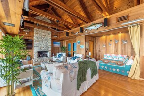 Interior acolhedor de cabana com lareira de pedra, vigas de madeira no teto, cama com tema náutico e uma grande planta em vaso.