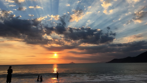 Solnedgang over vand med folk i vandet og på stranden.