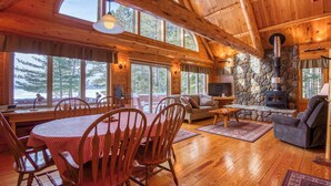 A cozy cabin interior with a dining area, seating, a fireplace, and large windows offering a view of the outdoors. - Two Winds - Beautiful newer home on Mooselookmeguntic Lake (Rangeley)
