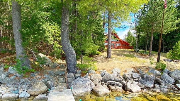 A wooden dock leading to a clear pond surrounded by rocks, with a red building in the background. - Two Winds - Beautiful newer home on Mooselookmeguntic Lake (Rangeley)
