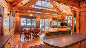 A wooden cabin interior with a dining area, kitchen, and large windows. - Two Winds - Beautiful newer home on Mooselookmeguntic Lake (Rangeley)