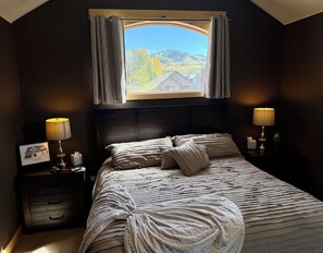 A bedroom with a large bed, two bedside lamps, a framed picture, and a window with curtains.