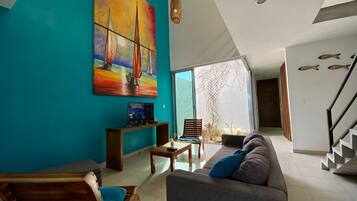 A modern living room with a teal wall, a large sailboat painting, a wooden chair, a gray sofa, and a staircase.