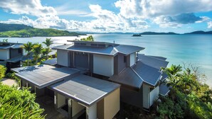 Um imóvel costeiro com vários edifícios, incluindo uma casa com telhado inclinado e uma garagem, rodeado por vegetação exuberante e com vista para um mar calmo e ilhas ao longe.