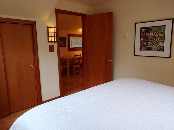 A bedroom with a large bed, wooden doors, a dining area with a table and chairs, and a framed artwork on the wall.