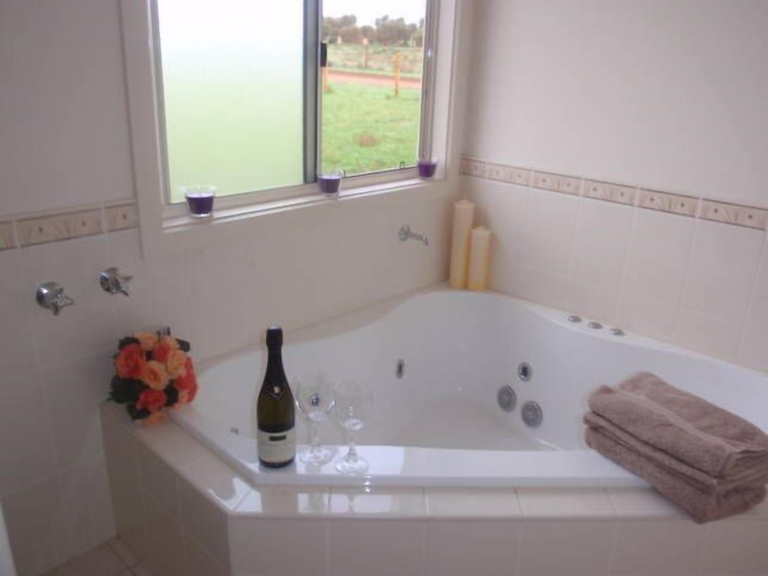 A bathroom with a large bathtub, a window with a view of a green field, and a bottle of wine on the edge of the tub.