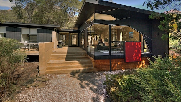 Une maison moderne dotée d’une grande baie vitrée, d’une terrasse en bois et d’un mur d’accent de couleur rouge.