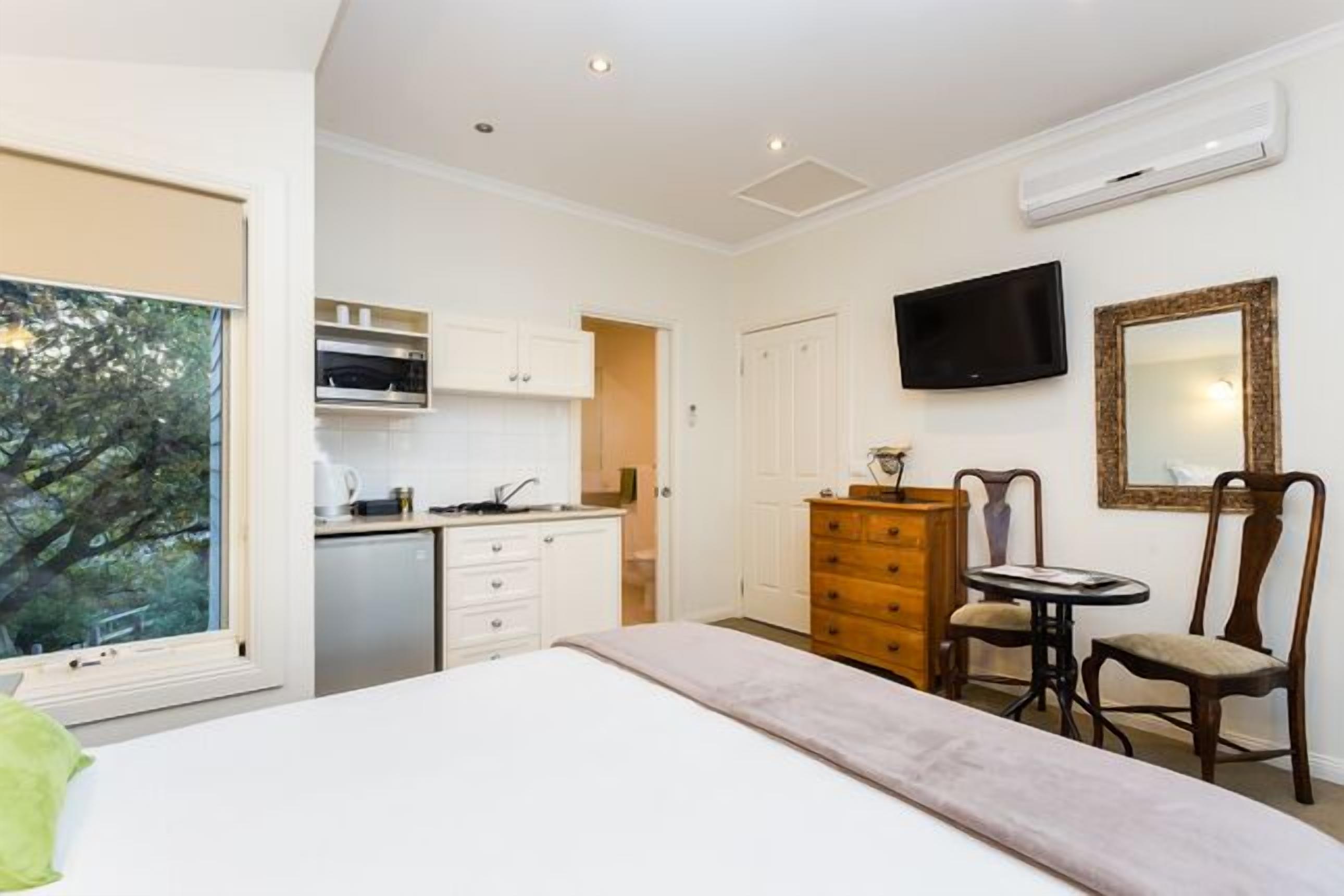 A hotel room with a bed, a small kitchenette, a TV, a mirror, and a window with a view of trees.