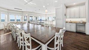 Een ruime eethoek met een grote houten tafel omringd door witte stoelen, een keuken met witte kasten en grote ramen met uitzicht.