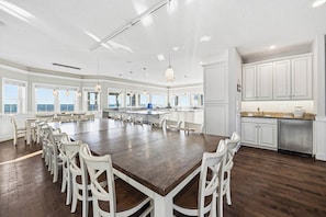 A spacious dining area with a large wooden table surrounded by white chairs, a kitchen with white cabinets, and large windows offering views. - Aquatic Dreams | Oceanfront | Private Pool, Hot Tub (Virginia Beach)