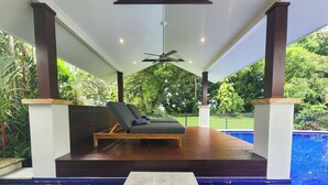 Un área de piscina techada con sillones reclinables, un ventilador de techo y vistas a la vegetación circundante.