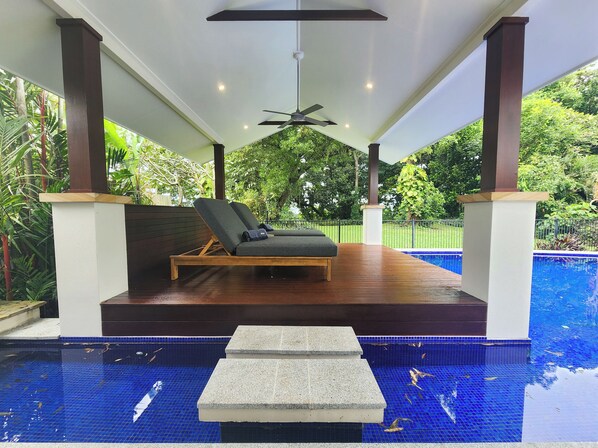 A covered pool area with lounge chairs, a ceiling fan, and a view of the surrounding greenery. - Pearl on Newell Beach 26 Rankin Street, Newell Beach QLD 4873 (Newell)