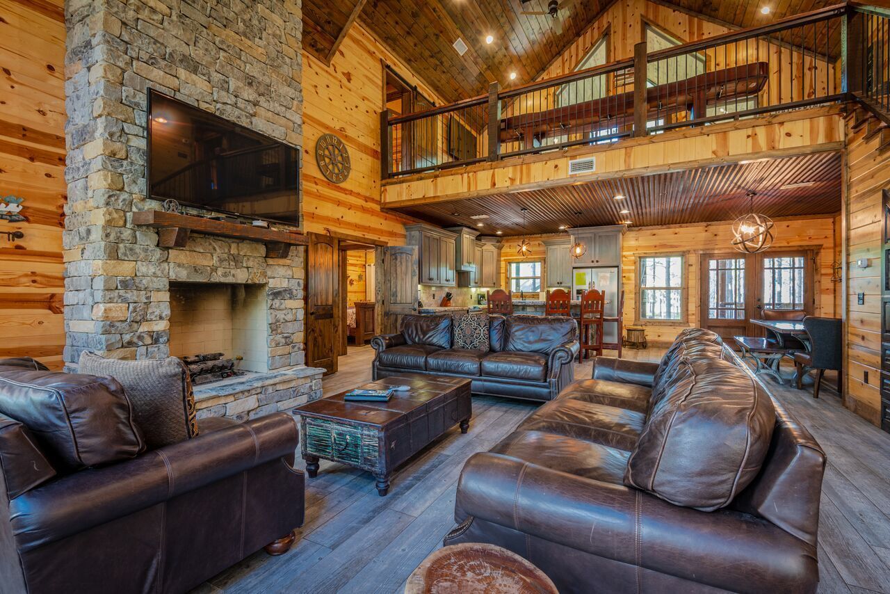 A spacious log cabin interior with a stone fireplace, leather sofas, and a balcony overlooking the yard.