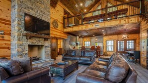 A spacious log cabin interior with a stone fireplace, leather sofas, and a balcony overlooking the yard. - Luxury Broken Bow Cabin w/ Private Pond | Sleeps 18 (broken bow)