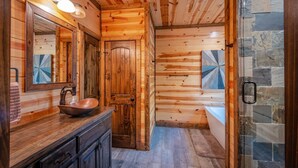 A wooden cabin bathroom with a large mirror, a wooden vanity, a stone shower enclosure, and a rustic wooden door. - Luxury Broken Bow Cabin w/ Private Pond | Sleeps 18 (broken bow)