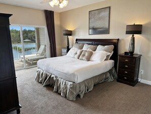 A bedroom with a large bed, a nightstand with a lamp, a dresser, and a view of a balcony with outdoor furniture.