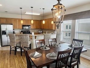 A modern kitchen with wooden cabinets, a stainless steel refrigerator, a microwave, and a coffee maker. There are wicker chairs around a wooden dining table with placemats.