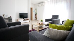 Ein modernes Wohnzimmer mit Fernseher, grauen Sofas, einem Couchtisch und einem Essbereich im Hintergrund.