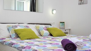 Ein ordentlich bezogenes Bett mit einer farbenfrohen, gemusterten Tagesdecke, zwei grünen Kissen und einem aufgerollten violetten Handtuch.