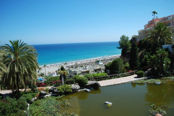 A coastal view with a beach, palm trees, and a building with balconies overlooking the sea.