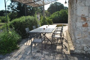 Eine Terrasse im Freien mit einem rechteckigen Tisch und mehreren Stühlen, umgeben von Grünpflanzen und einer Steinsäule.
