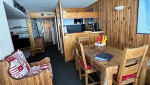 Quarto compacto com mesa de jantar de madeira, cadeiras, pequena cozinha e um canto de estar acolhedor com almofada estampada.