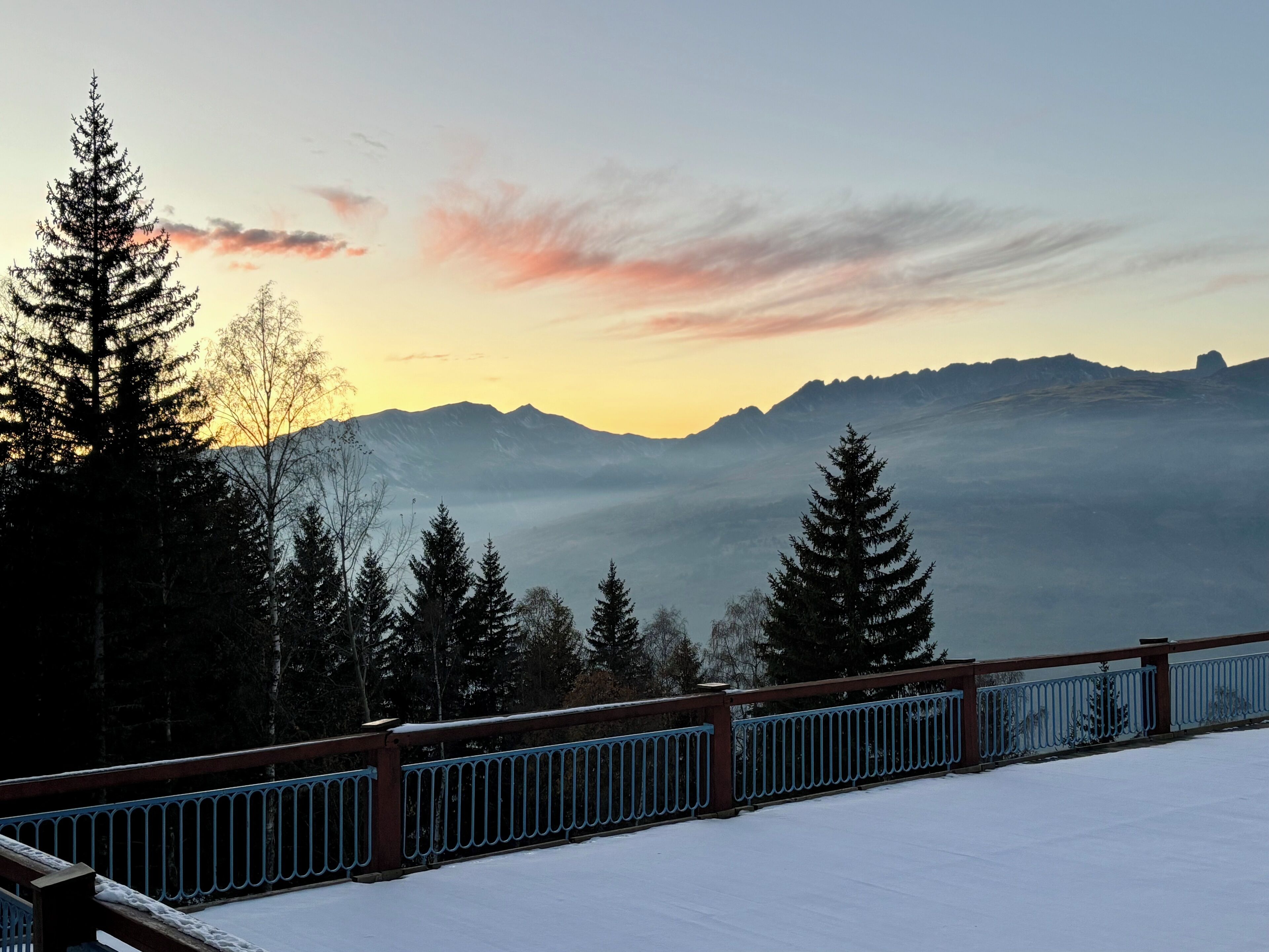 A snow-covered deck with a metal railing, overlooking a mountainous landscape with trees and a sunset.