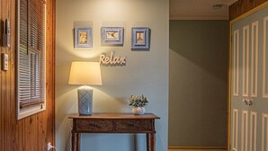 Uma secretária de madeira com uma lâmpada e objetos decorativos, uma parede com quadros e um letreiro com a palavra 'Relax', e uma porta com painéis verticais.
