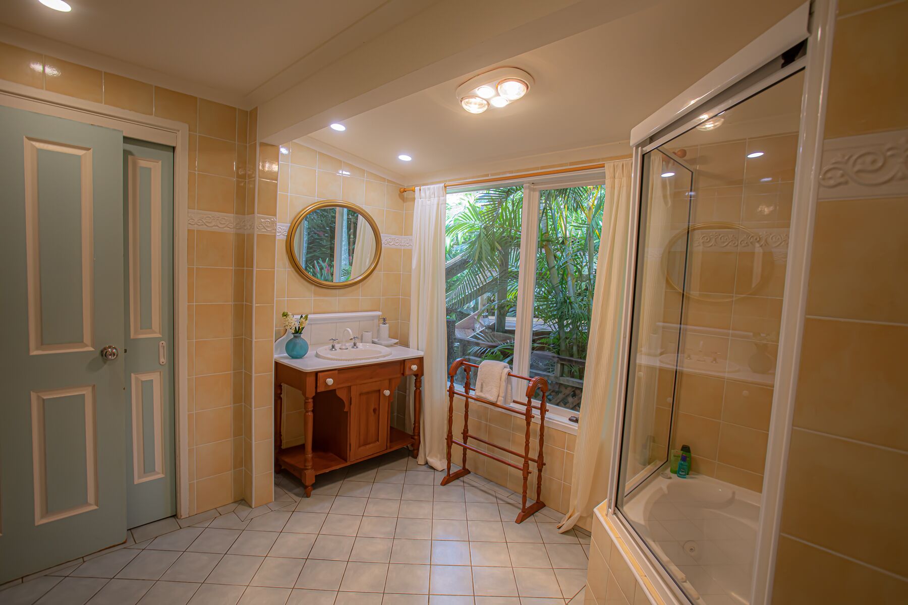 A bathroom with a wooden vanity, a round mirror, a shower with a glass door, and a window with a view of greenery.