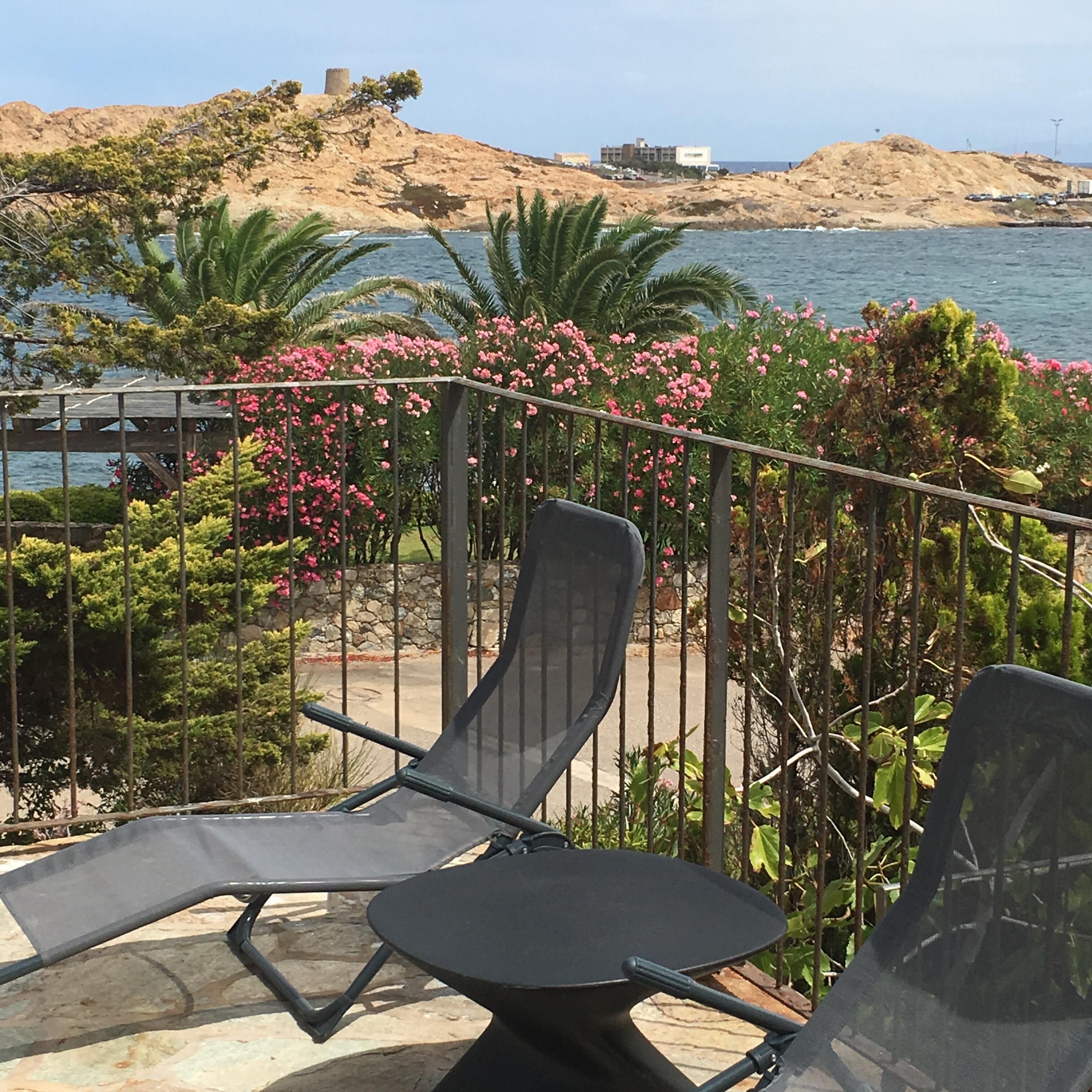 Un balcon avec deux fauteuils et une table, donnant sur une zone côtière caractérisée par un terrain rocheux et une étendue d’eau.