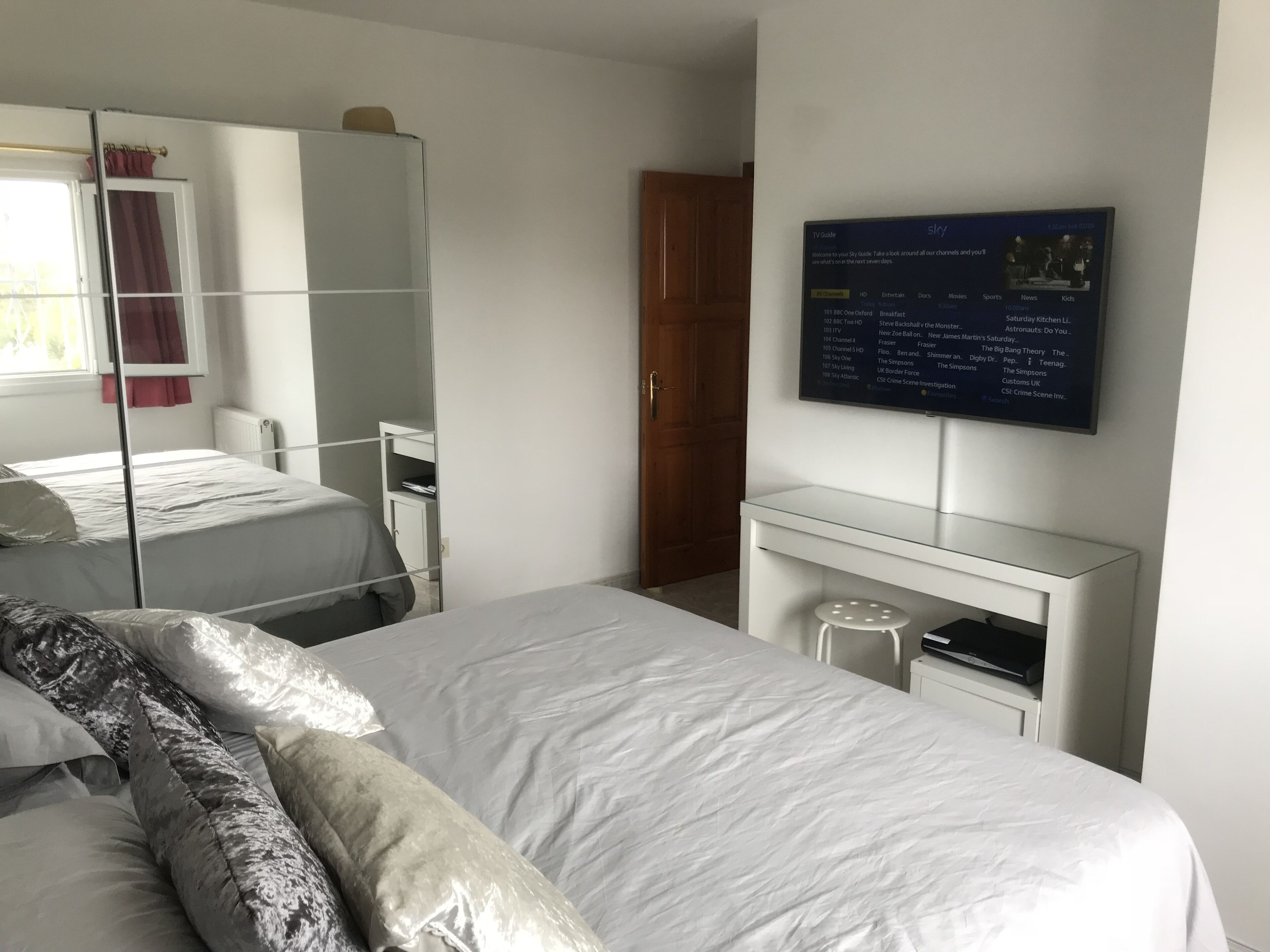 A bedroom with a bed, a wardrobe, a desk, and a TV mounted on the wall displaying a TV guide.