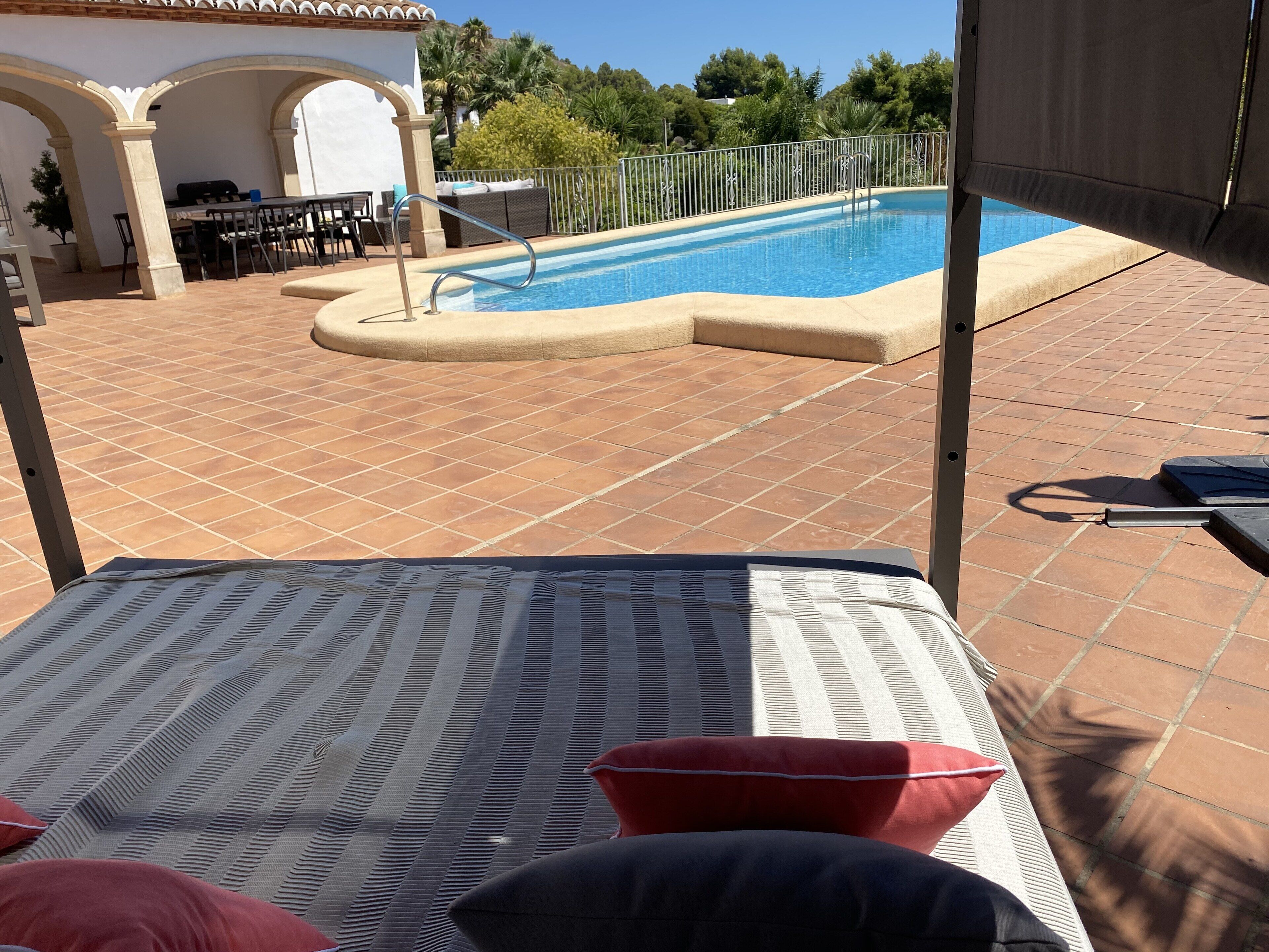 A pool area with a lounge chair and cushions, a dining table with chairs, and a patio with tiled flooring.