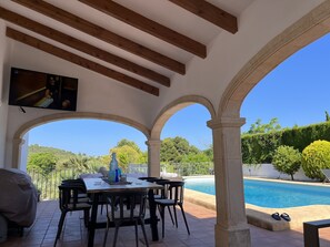 A covered outdoor area with a pool, dining table, chairs, and a TV mounted on the wall.