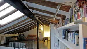 Ein geräumiger Dachboden mit freiliegenden Holzbalken, einer Treppe, einer Couch und einem Bücherregal voller Bücher.