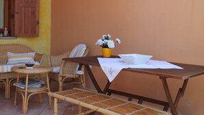 Un conjunto de mobiliario de mimbre con una mesa y sillas, una pequeña mesa redonda con un bol, y una maceta amarilla con flores sobre un mantel.