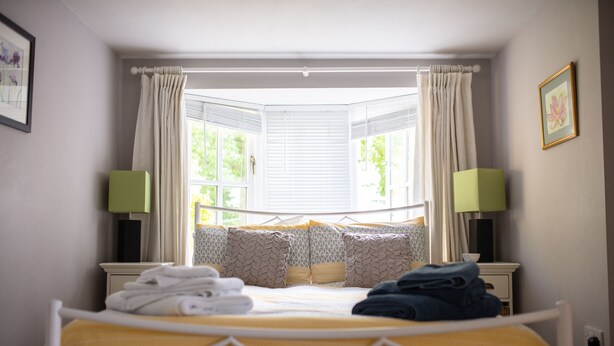 Ein ordentlich gemachtes Bett mit gelber Tagesdecke, weißen Kissen und gefalteten Handtüchern. Zwei Nachttische mit grünen Lampenschirmen, ein Fenster mit weißen Vorhängen und ein gerahmtes Bild an der Wand sind ebenfalls zu sehen.