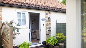 Ein charmantes Cottage mit weißer Fassade, einem Ziegeldach und einer Holztür. Auf der Veranda stehen Blumentöpfe und ein kleiner Tritt führt zum Eingang.