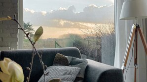Ein modernes Wohnzimmer mit einem grauen Sofa, einer Stehlampe und Blick auf einen bewölkten Himmel durch ein großes Fenster.